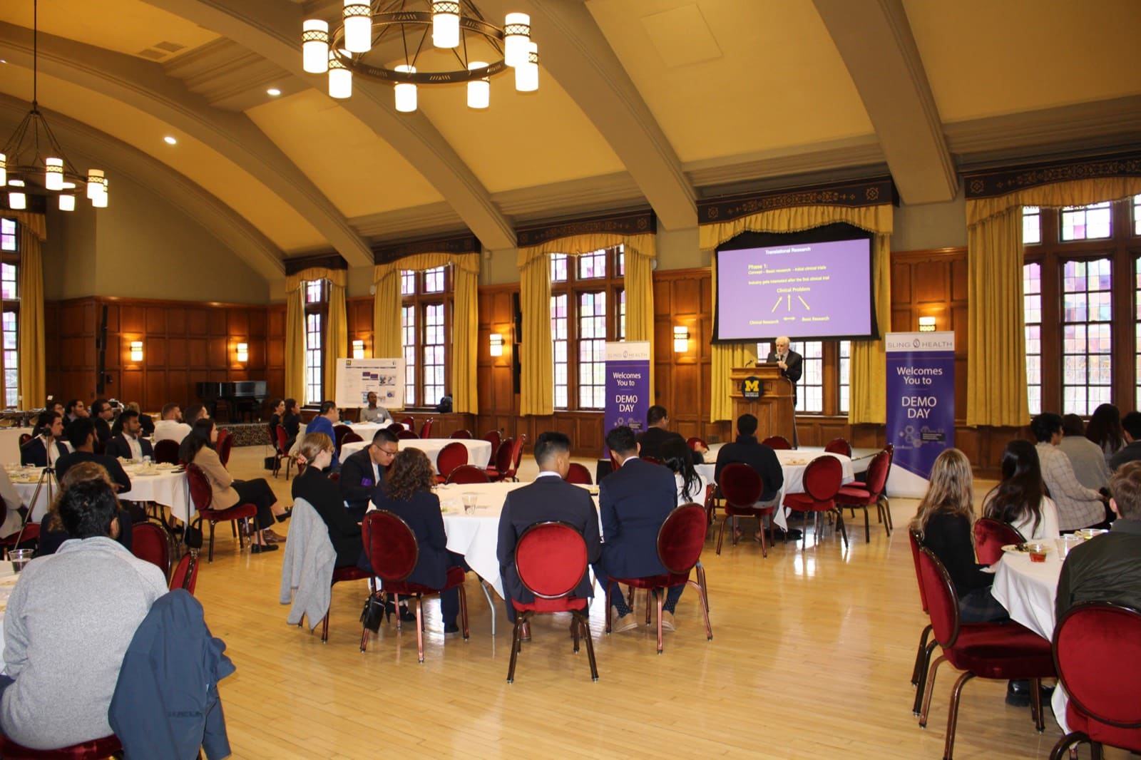 A Sling Health Demo Day hosted in a university hall, tables of attendees facing the stage.